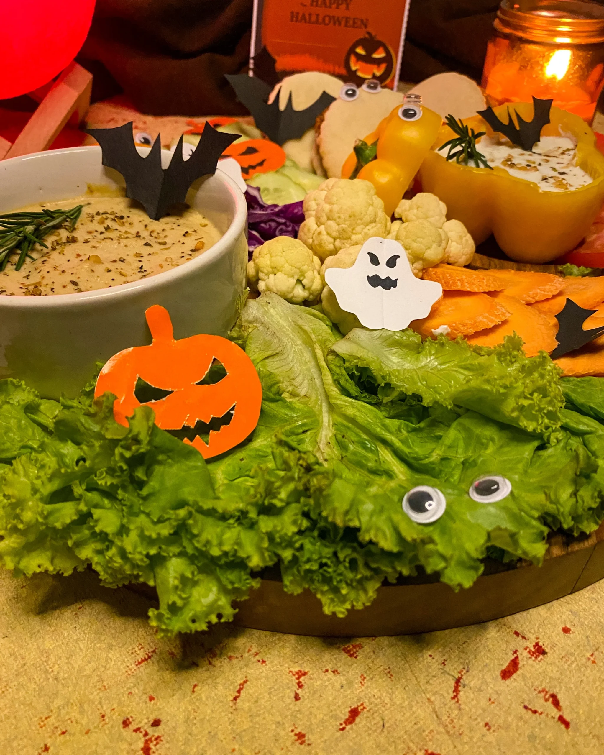 Halloween crudité platter with spooky decorations featuring fresh vegetables, Mediterranean hummus, yellow pepper tzatziki bowl, and festive bat ghost cutouts on wooden serving board