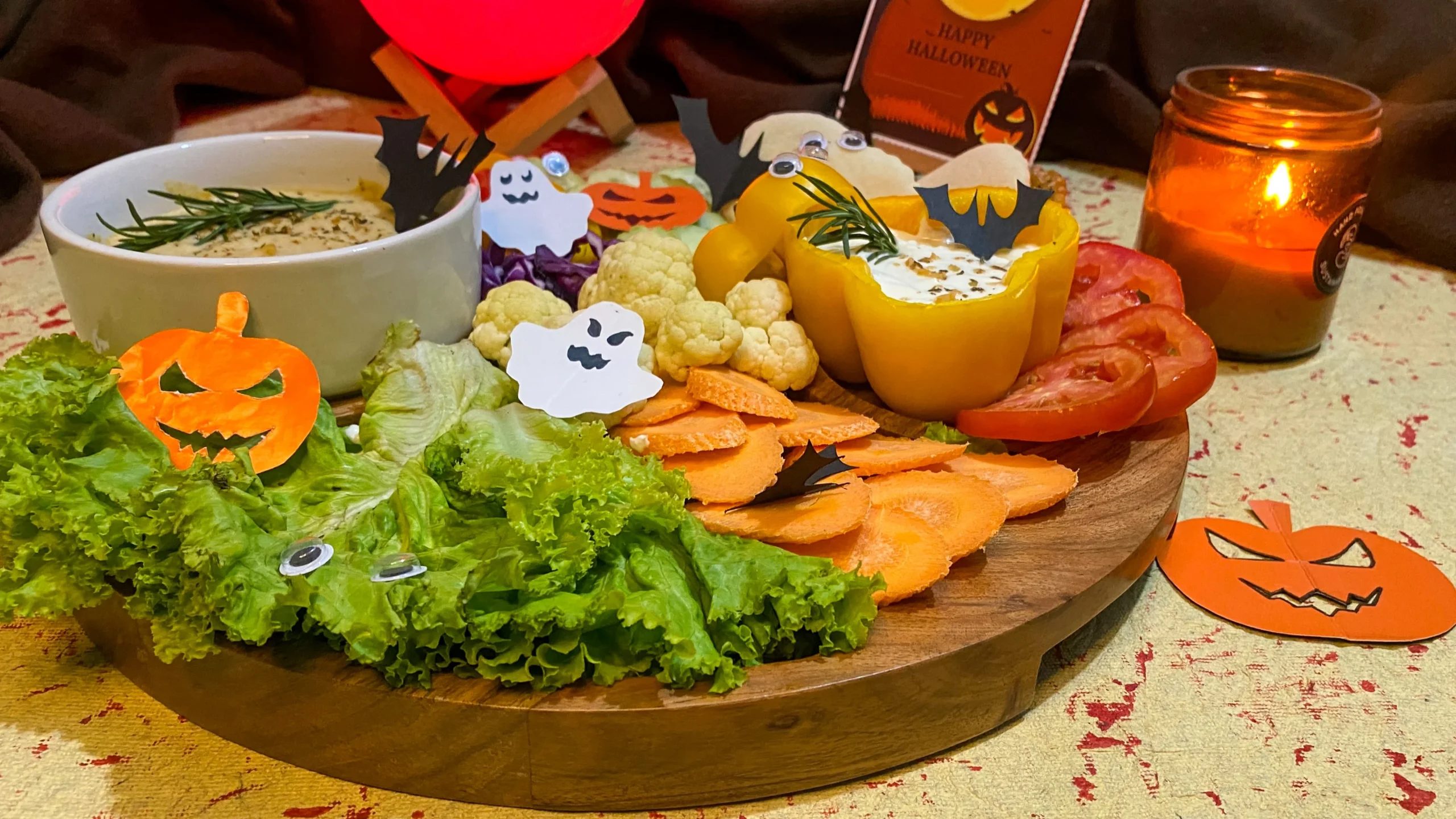 Healthy Halloween veggie platter on wooden chip and dip board with Mediterranean dips, cauliflower, carrots, tomatoes, bell peppers, and spooky pumpkin ghost decorations with candle ambiance