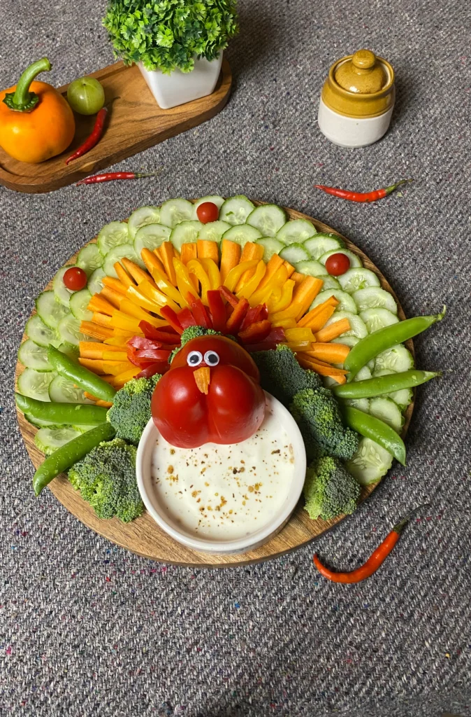 Festive Thanksgiving turkey veggie tray with colorful cucumber, carrot, broccoli, snap peas, and cherry tomatoes arranged around garlic dill yogurt dip in wooden platter