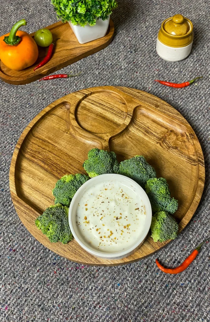 Empty rustic wooden serving platter on textured woven tabletop next to small ceramic pot, orange bell pepper, and green decorative plant